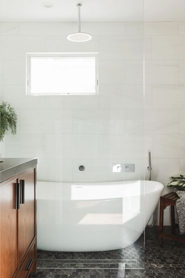 Freestanding white tub with a overhead rain shower, white walls, black hex tile flooring wood vanity