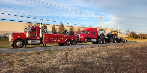 Red tow truck hauling two trucks on a trailer under a blue sky.