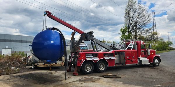 Red tow truck lifting a large blue cylindrical tank outdoors.