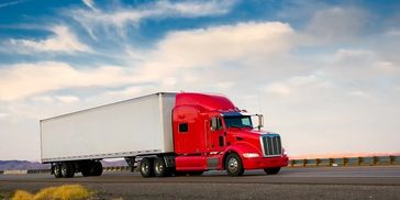 A red semi-truck driving on a highway under a partly cloudy sky.