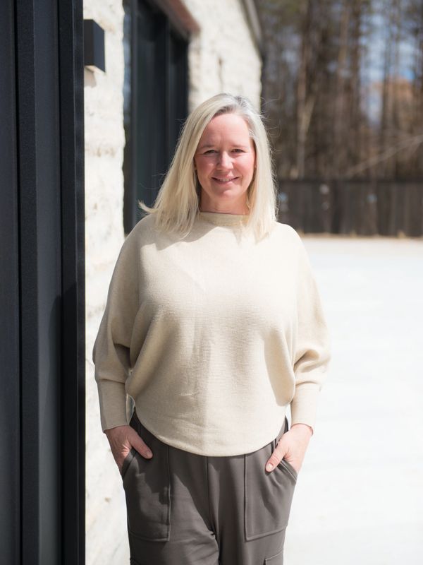 Woman in beige sweater and gray pants standing outside near a building.