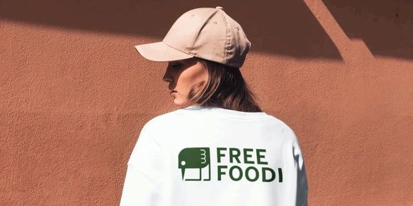 Person wearing a white shirt with "FREE FOOD!" printed on the back, standing against a brown wall.