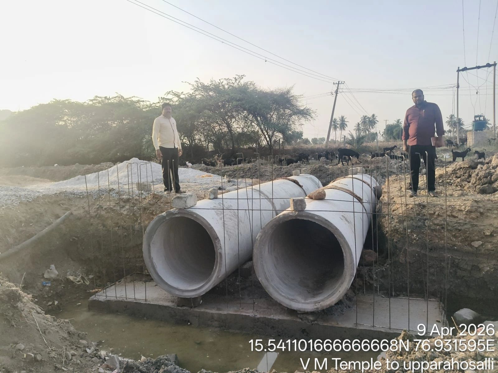 Two men stand beside large concrete drainage pipes at a construction site.