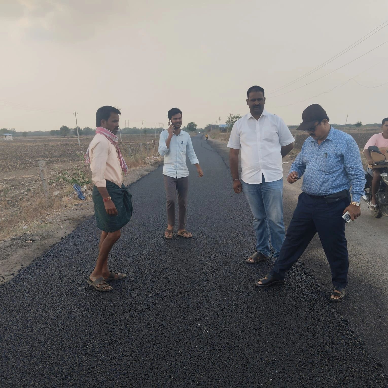 Four men standing on a newly paved rural road, with one talking on the phone.