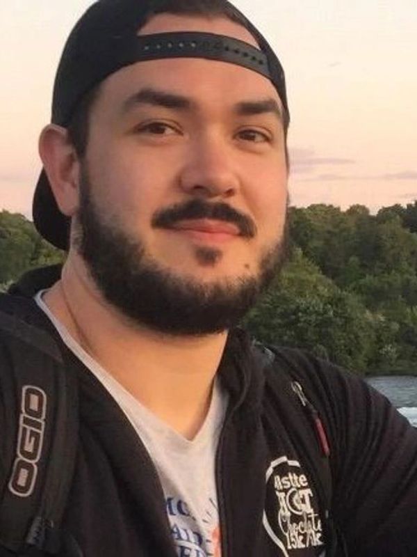 Bearded man wearing a backward cap and backpack outdoors near trees.