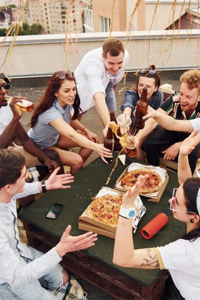 Group of friends toasting with drinks around pizza on a rooftop.