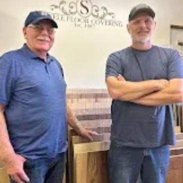 Two men standing in front of a display of wood flooring samples.