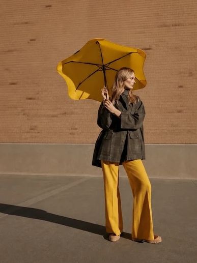Woman with yellow umbrella and pants standing against a brick wall.
