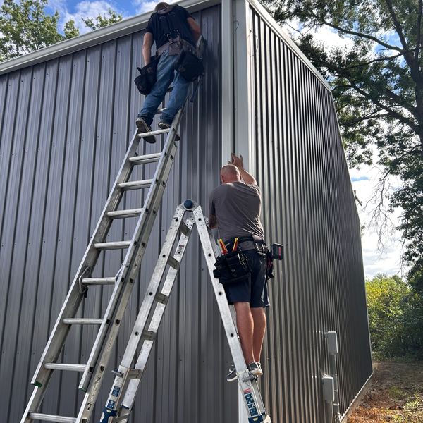 Two workers on ladders fixing a metal building exterior under a blue sky.