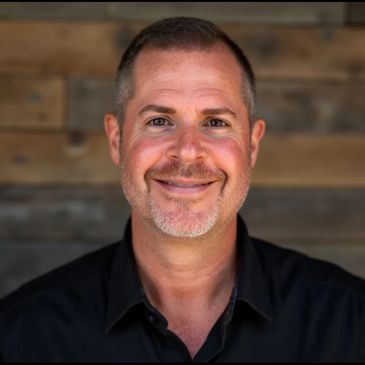 Smiling middle-aged man with short hair and a goatee, wearing a black shirt against a wooden background.