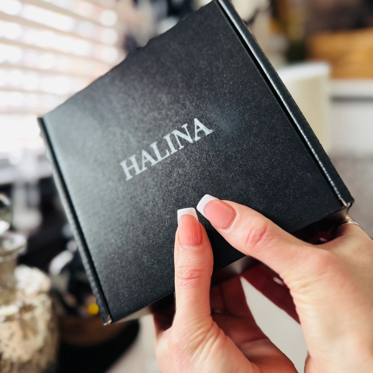 Hand holding a black box labeled 'HALINA' with French manicure nails.