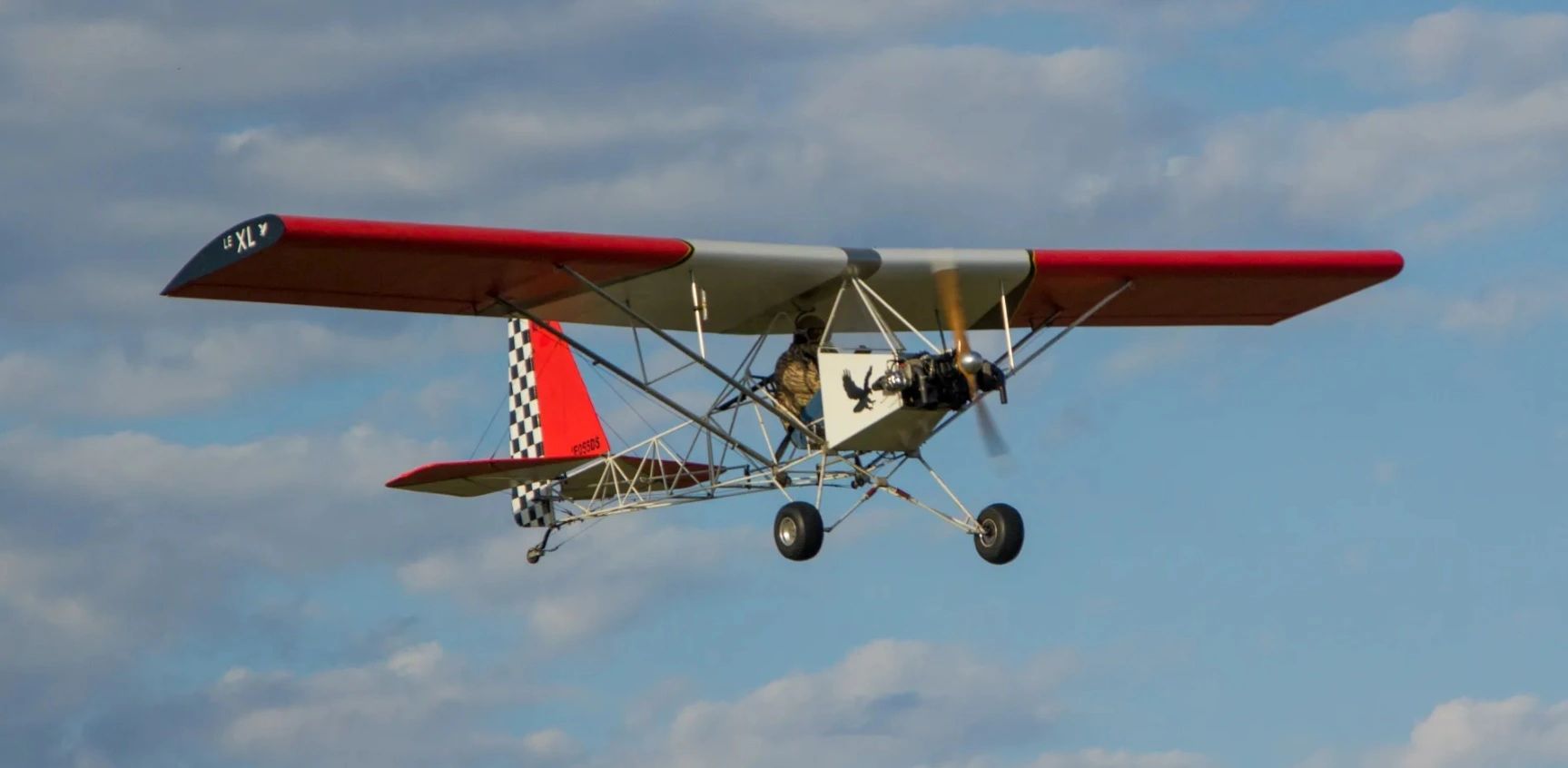 A lightweight, single-engine ultralight aircraft flying under a partly cloudy sky.