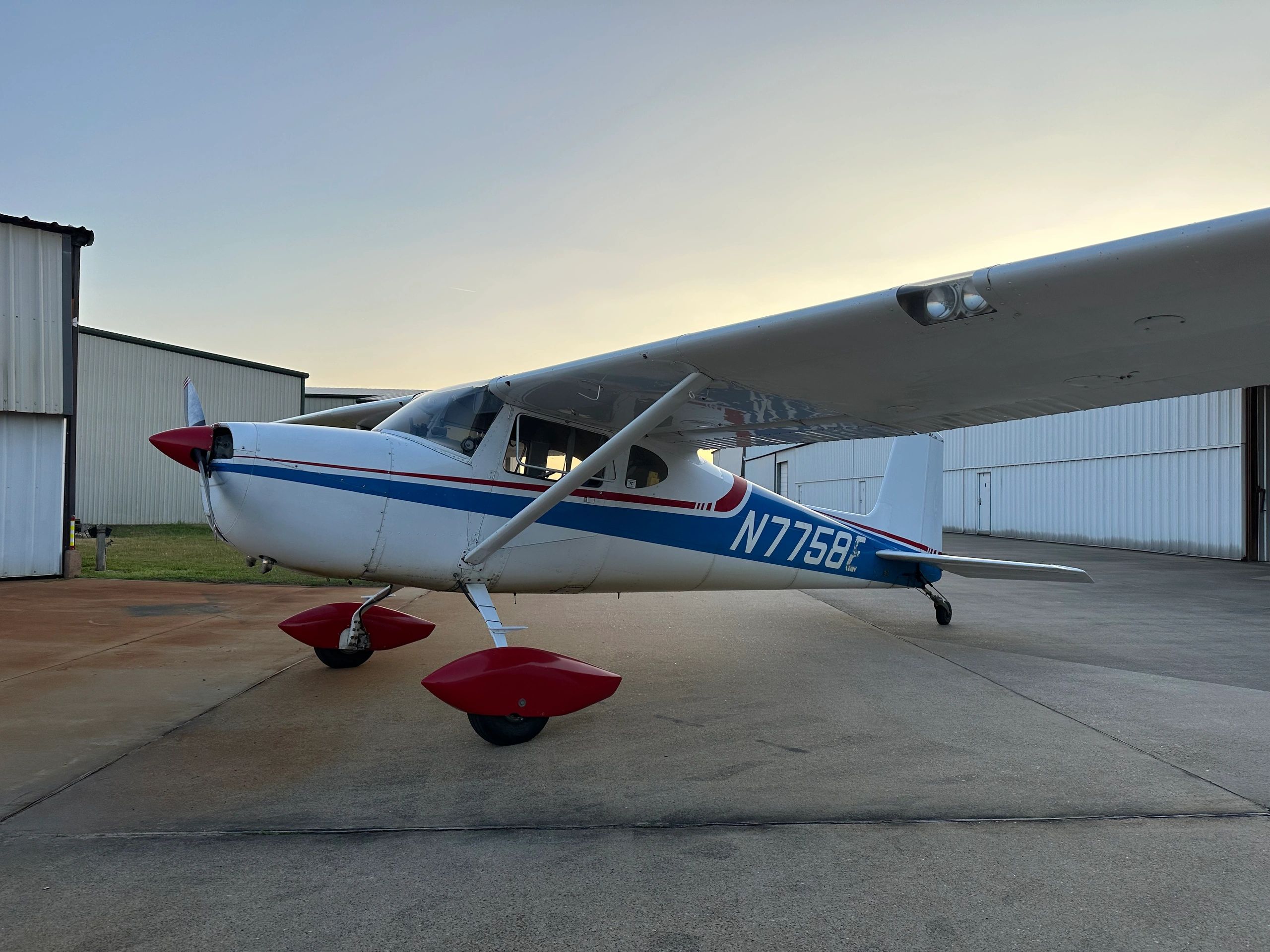 Small white and blue single-engine airplane parked on concrete near hangars.