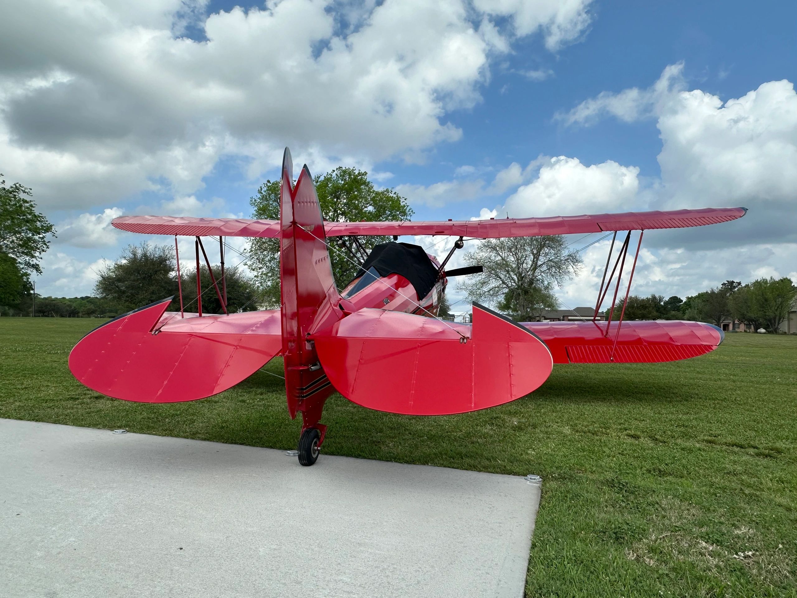 Rear view of a red vintage biplane on a grassy field under a cloudy sky.