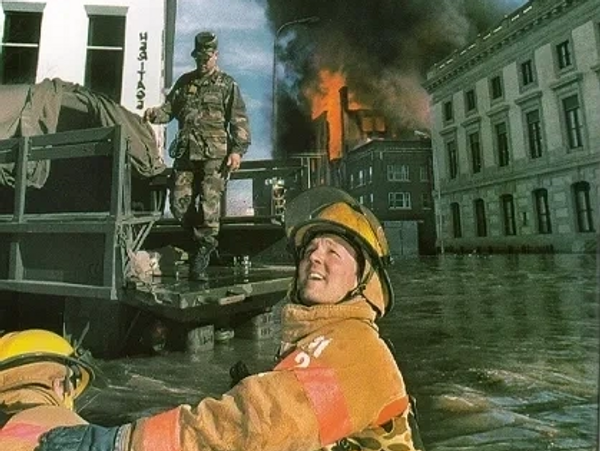 Two firemen in a flooded city street with a fire in the background.