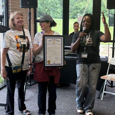 Three women at an indoor event, one holding a framed certificate, another speaking into a microphone.