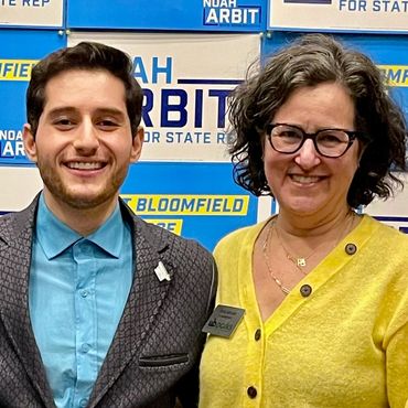 Two people smiling in front of political campaign signs for Noah Arbit.