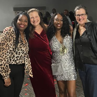 Four women smiling together at an indoor event, dressed in stylish outfits.