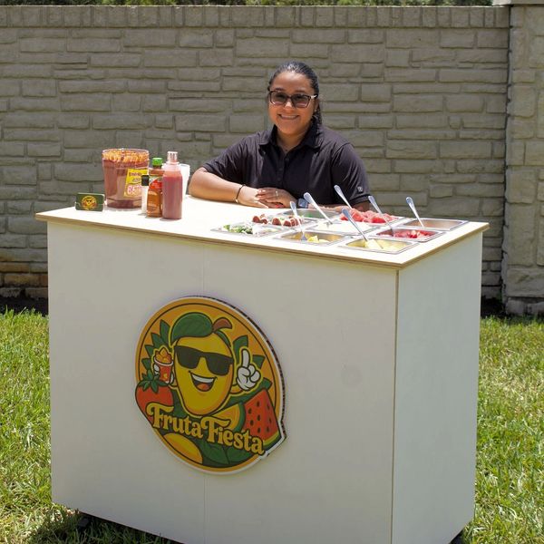 A woman smiles behind a Fruta Fiesta fruit stand with fresh fruit and sauces outdoors.