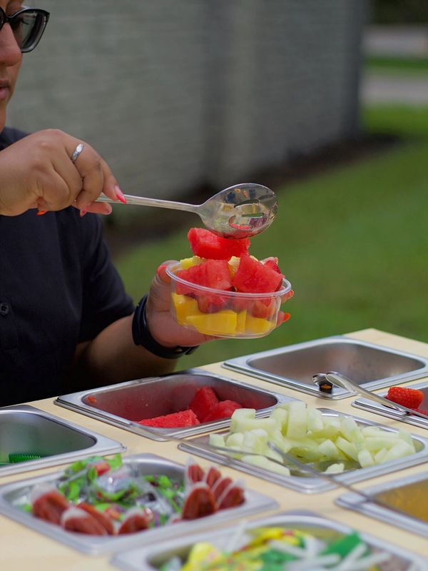 Person serving fresh fruit salad from a buffet table outdoors.