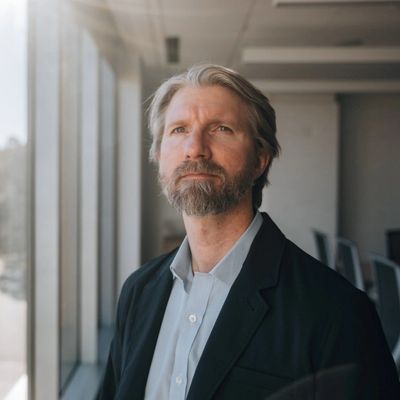 A thoughtful man with gray hair and beard stands by a window in a modern office.