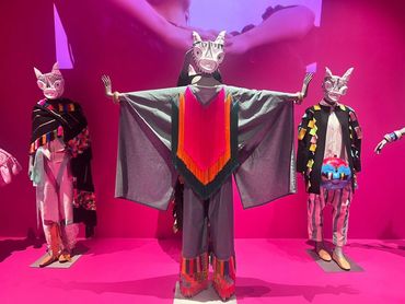 Three mannequins wearing colorful animal masks and vibrant costumes on a pink stage.