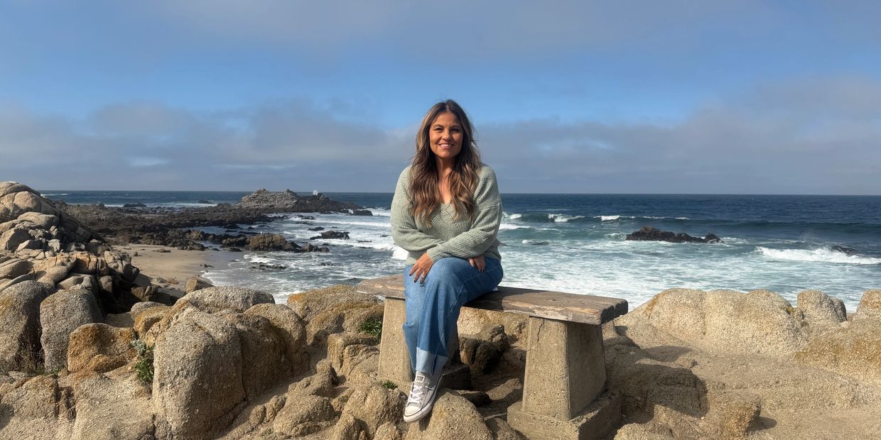 A woman sitting on a bench by rocky ocean shore under blue sky.