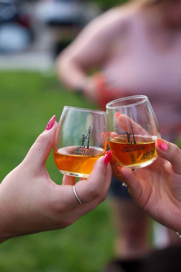 Two hands clinking glasses with amber liquid outdoors.