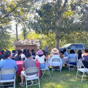 Group gathered outdoors listening to a speaker under trees.