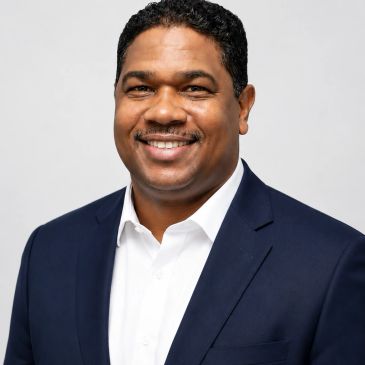 Smiling man in a navy blue suit and white shirt against a plain background.