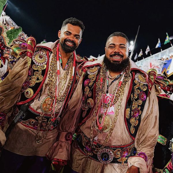 Two men smiling in ornate, colorful traditional costumes with gold chains, at a nighttime event.