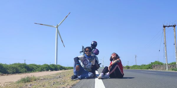Three friends posing on an open road with a wind turbine in the background under a clear blue sky.