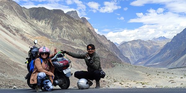 Two motorcyclists posing with their bike in a mountainous desert landscape.