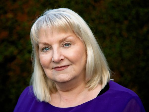 Smiling older woman with blonde hair wearing a purple top against a dark background.