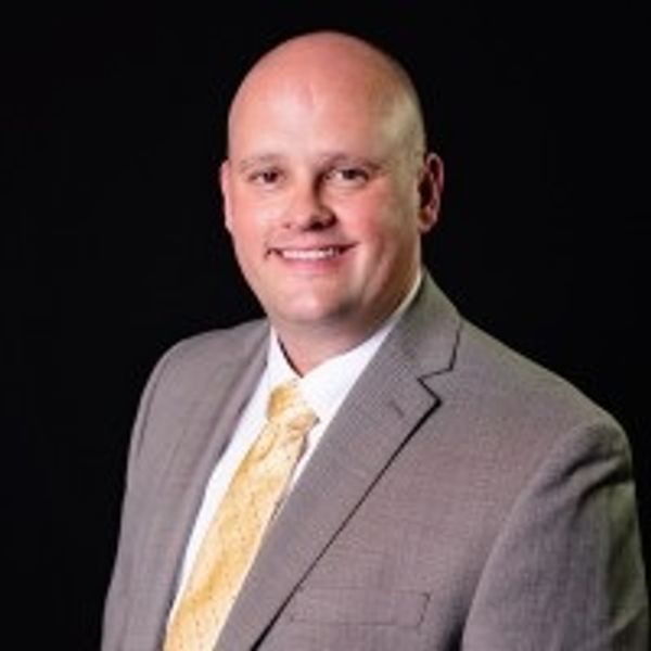Smiling bald man in a gray suit and yellow tie against a black background.