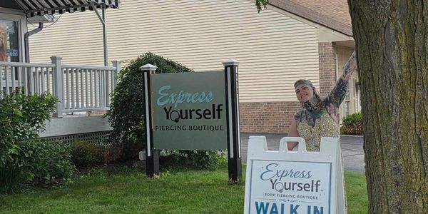 Person poses happily by 'Express Yourself Piercing Boutique' signs.