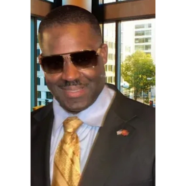 Man in suit with gold tie and sunglasses smiling indoors.