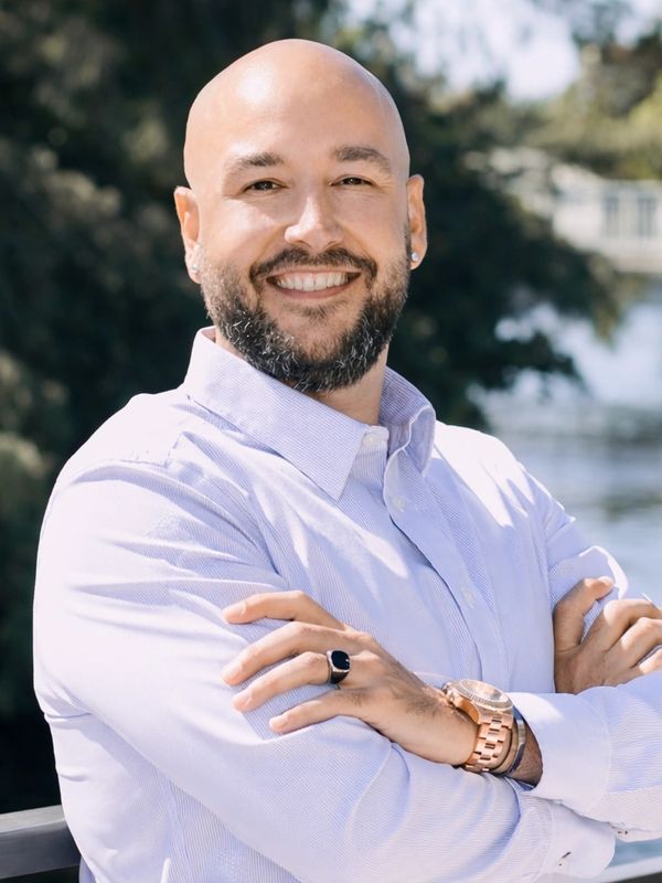 Smiling bald man with beard, wearing a light shirt and jewelry, standing outdoors.