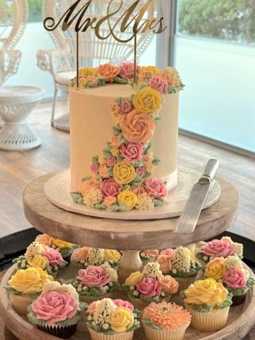 Elegant wedding cake and cupcakes adorned with pastel floral decorations and a "Mr &amp; Mrs" topper.