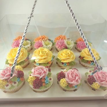 Assorted cupcakes decorated with colorful buttercream flowers in a clear box.