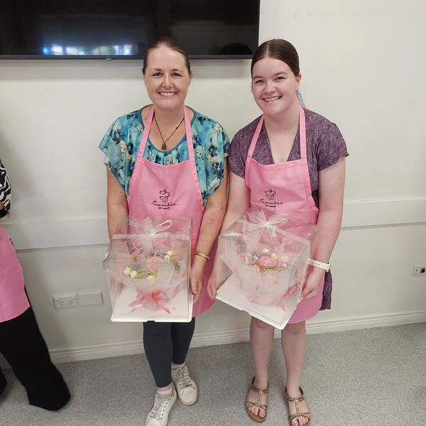 Two women in pink aprons holding cakes wrapped in clear boxes.