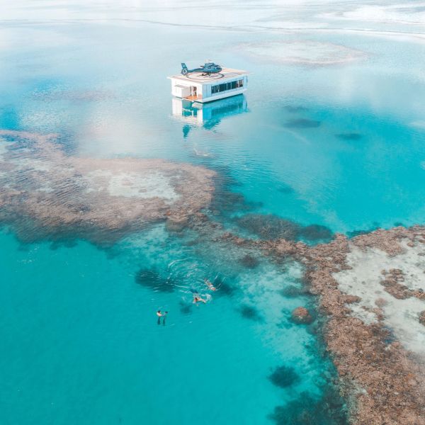 A floating house with a helicopter on top in clear blue water above coral reefs.