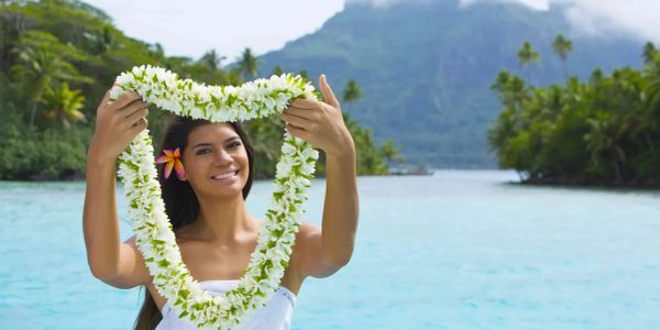 Woman holding a flower lei by the water with mountains in background.