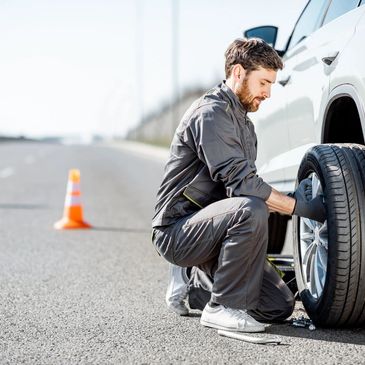 A man changing a car tire on the side of the road with a safety cone behind.