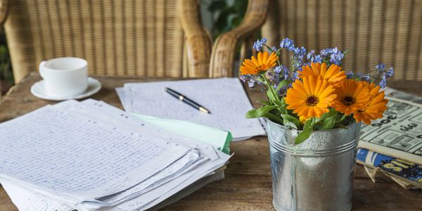 A rustic table with handwritten papers, a pen, a cup, and a metal vase of orange flowers.