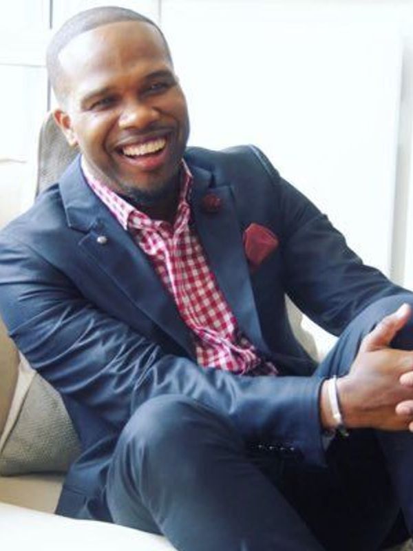 A man in a navy suit and red checkered shirt smiling broadly.