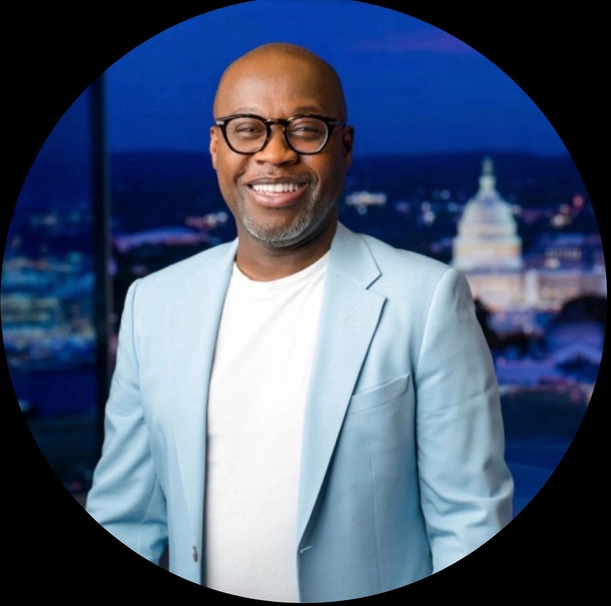 Smiling man in glasses and light blue blazer with city nightscape background.