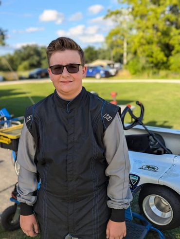 Teen in a racing suit stands beside a go-kart on a sunny day.