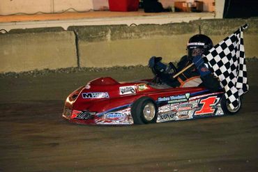 A go-kart driver holding a checkered flag, celebrating a win on a dirt track.