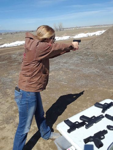 Woman aiming a pistol at an outdoor shooting range with several guns on a table.